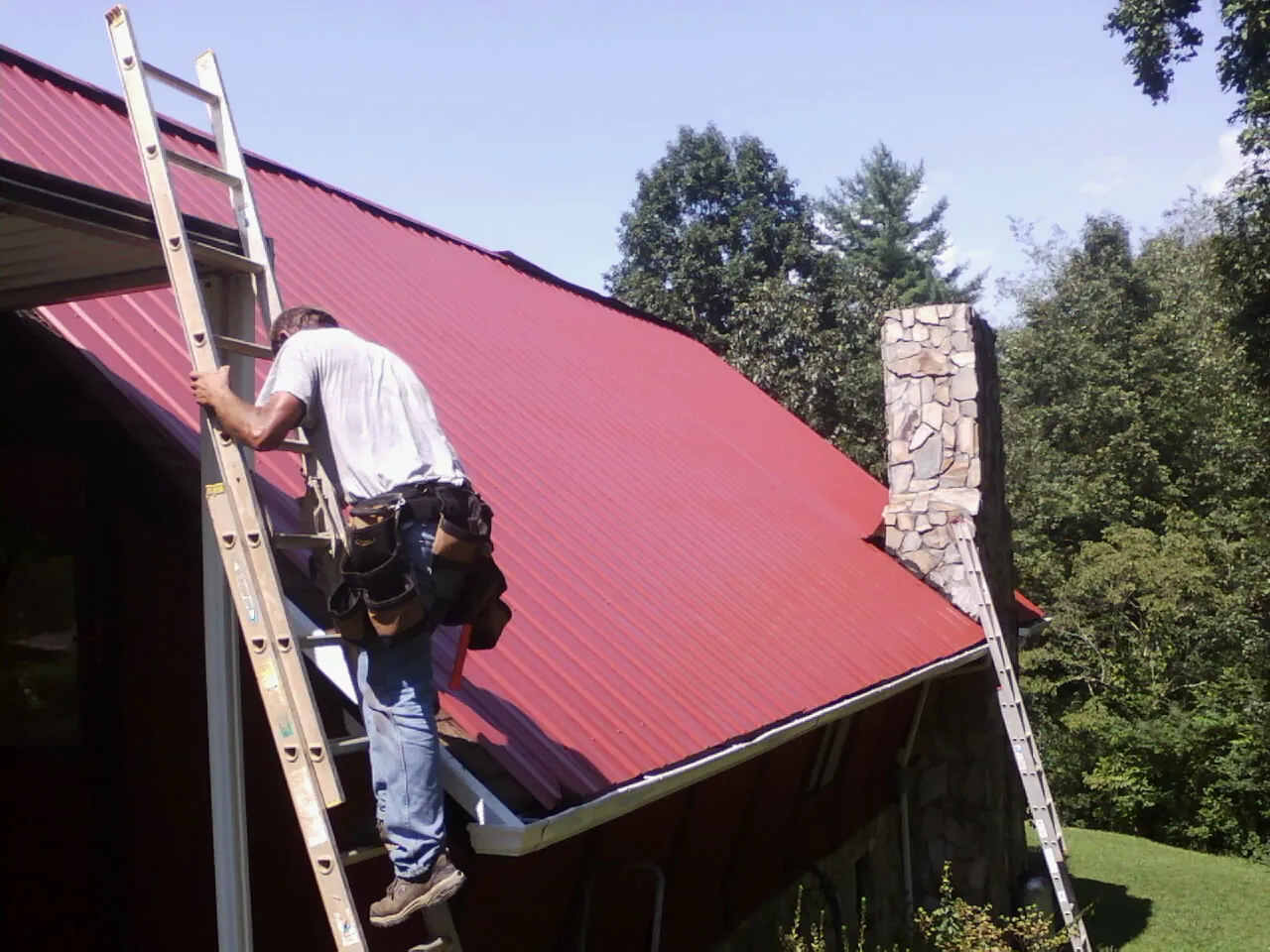 Roofer on a ladder beside a red metal roof and stone chimney — Soffit & Fascia Repair by Roofing Brothers in Selbyville